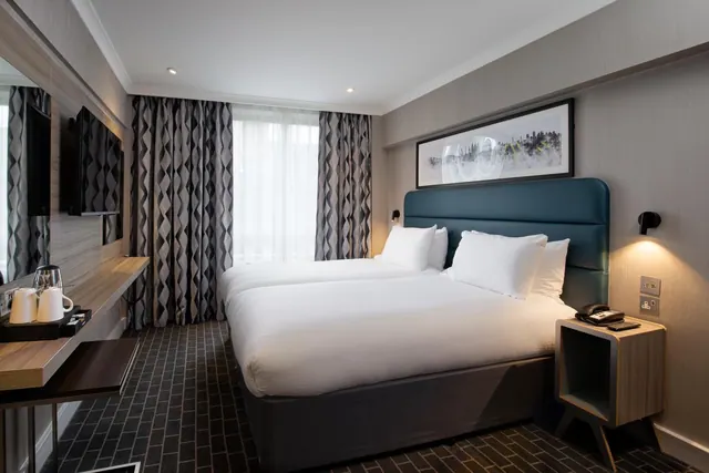 A hotel bedroom with two beds under a dark teal headboard, a framed print above them, a wall-mounted television and a kettle on a tray at the left.