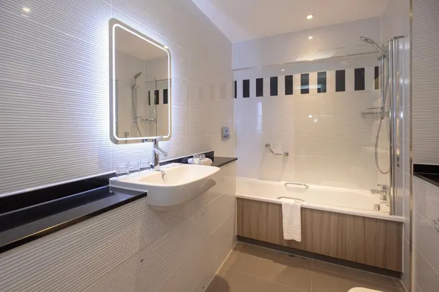 A bathroom with a wall-mounted sink below a backlit mirror, and a bath with a glass shower screen and grab rail.
