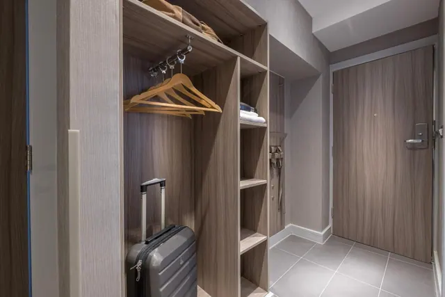 An entrance area of a hotel room with an open wardrobe holding wooden hangers, a grey suitcase on the floor, and a wooden entry door with a keycard lock.