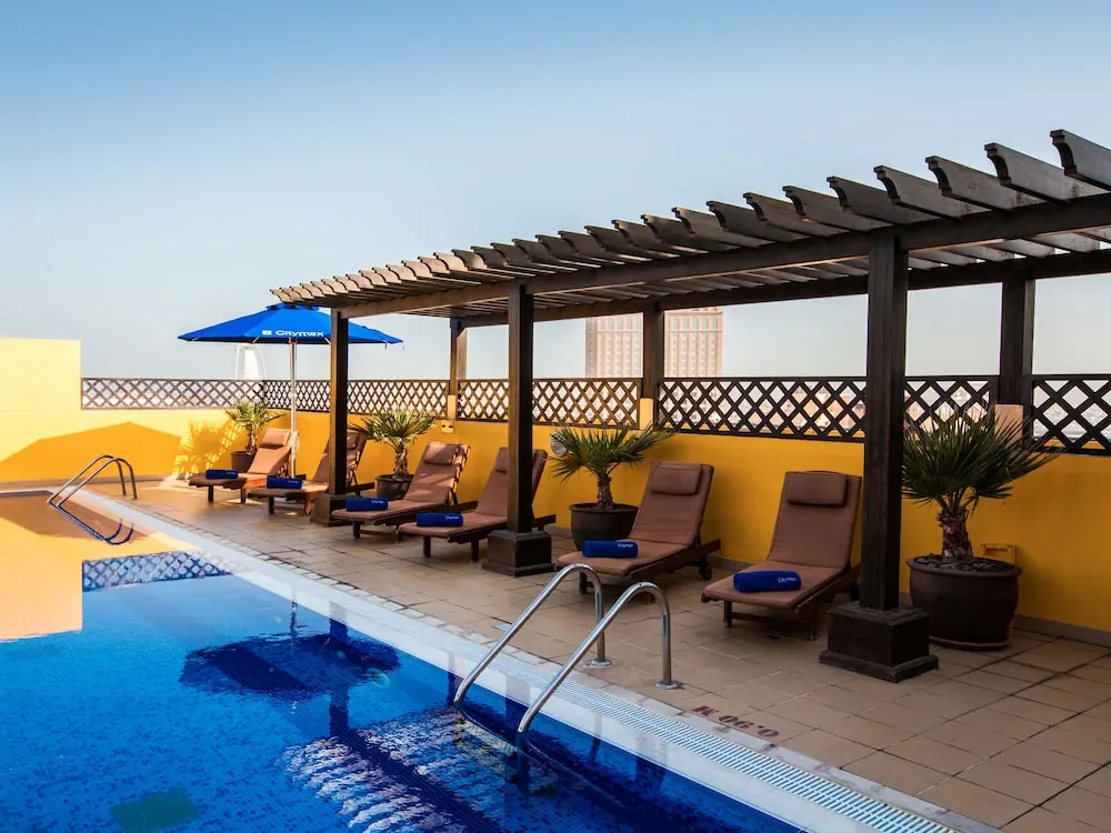 A rooftop swimming pool with sun loungers, blue towels and a blue umbrella beneath a wooden pergola.