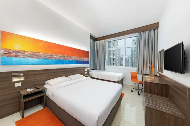 A hotel room with a double bed, a single bed behind it, an orange desk chair, a wall-mounted TV and a window overlooking residential towers.