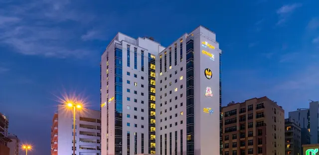 Exterior of a white high-rise hotel with illuminated CityMax Hotel signage at dusk, with street lamps and neighbouring buildings beside it.