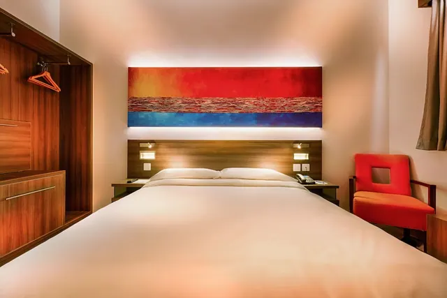 A bedroom with a double bed, a red armchair, a wooden wardrobe and a large abstract painting above the headboard.