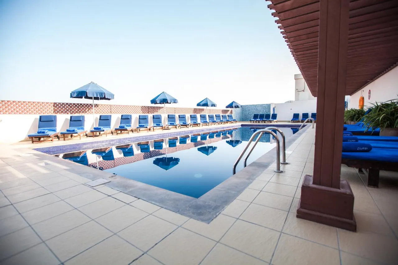 A rooftop swimming pool with blue sun loungers and blue parasols along its edge under a clear sky.
