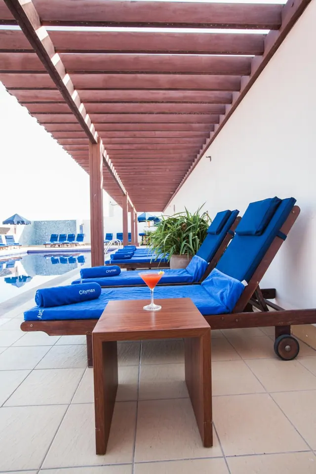 Blue-cushioned sun loungers and a cocktail on a wooden side table sit under a timber pergola beside a pool.