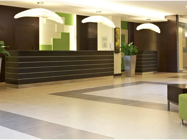 A hotel lobby with a long dark reception desk, three white pendant lights, green accent wall panels, potted plants and a green upholstered seat at the right edge.