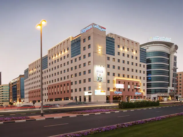 Exterior view of the Citymax hotel building at dusk, with illuminated windows and signage, a street lamp, and a road lined with flowering beds in the foreground.