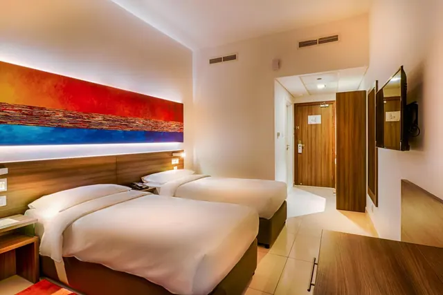 A hotel bedroom with two single beds, a colourful wall painting above the headboard, a wall-mounted television and a wooden desk in the foreground.