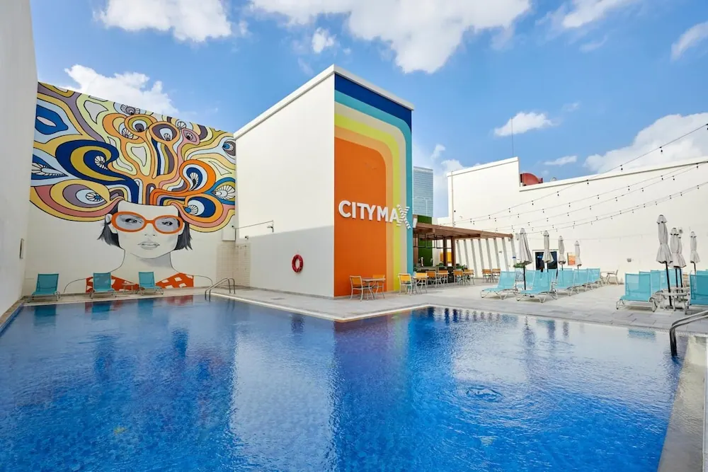 An outdoor pool with turquoise loungers, closed umbrellas and string lights, beside a wall with a colourful mural of a woman in orange glasses and a building with the words CITYMAX.