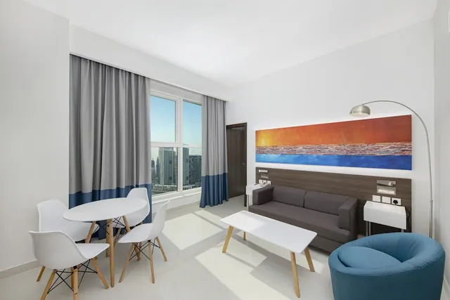 A hotel room living area with a grey sofa, a round table with white chairs, a large painted sunset artwork, and a window looking out over city towers.