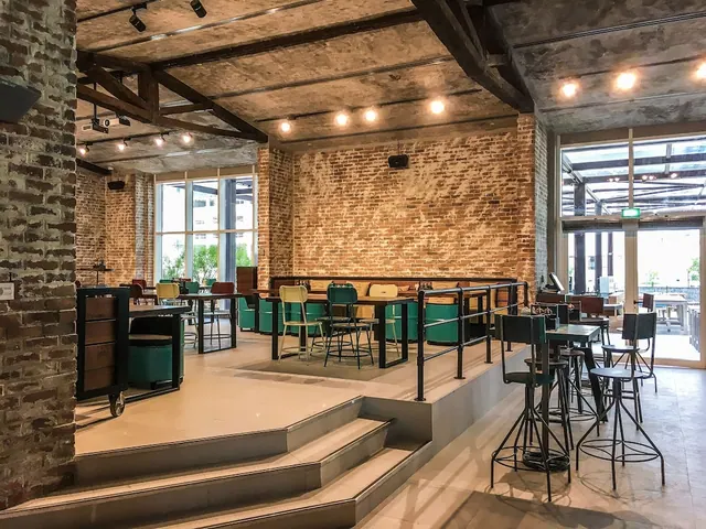 A restaurant dining area with exposed brick walls, wooden ceiling beams, green-upholstered booth seating, high bar tables and large glass windows looking out onto greenery.