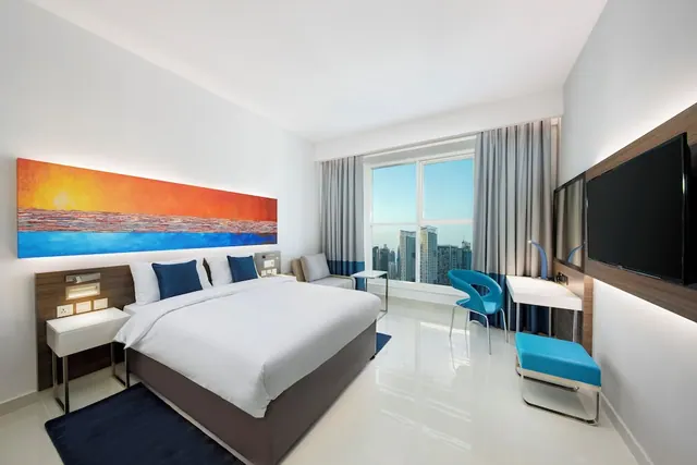A bedroom with a made king bed, a wall-mounted television, a desk with a blue chair, and a window overlooking high-rise towers.