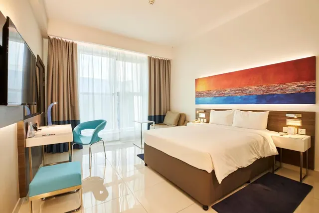 A hotel bedroom with a king bed, a turquoise chair at a white desk, an armchair by the sheer-curtained window, a wall-mounted television, and a large abstract painting above the bed.