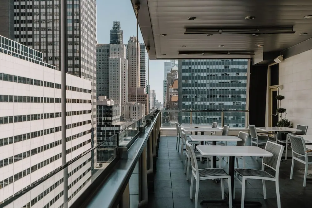 An outdoor terrace with white tables and chairs and a glass railing overlooks a cluster of tall city skyscrapers under a clear sky.