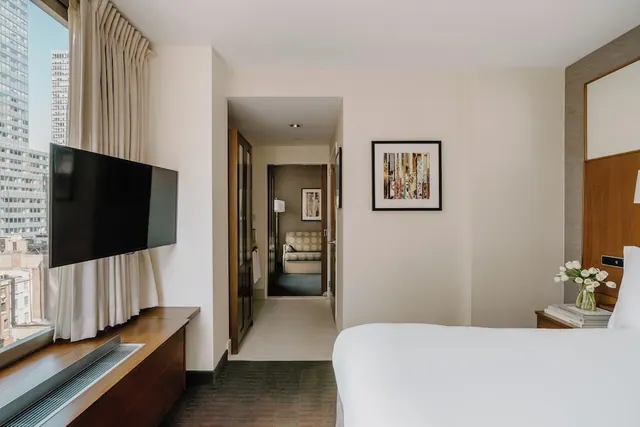 A bedroom with a made white bed, a wall-mounted television, and tall city buildings visible through the window, with a corridor leading to a seating area.