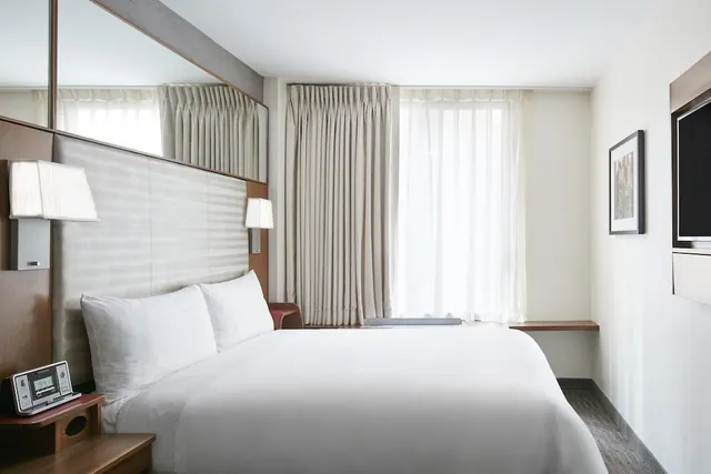 A bedroom with a made white bed, a wall-mounted television, a clock radio on the bedside table and sheer curtains over a window.