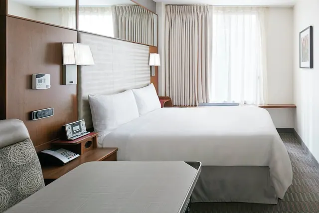 A hotel room with a large bed dressed in white linens, a wooden headboard wall with wall lamps and a clock radio, and an upholstered armchair in the foreground beside a curtained window.