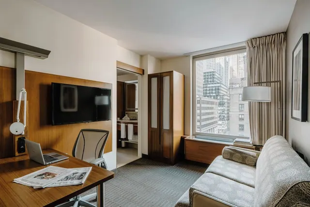 A hotel room with a wall-mounted television, a wooden desk with a laptop and newspapers, a sofa, and a window overlooking high-rise buildings.