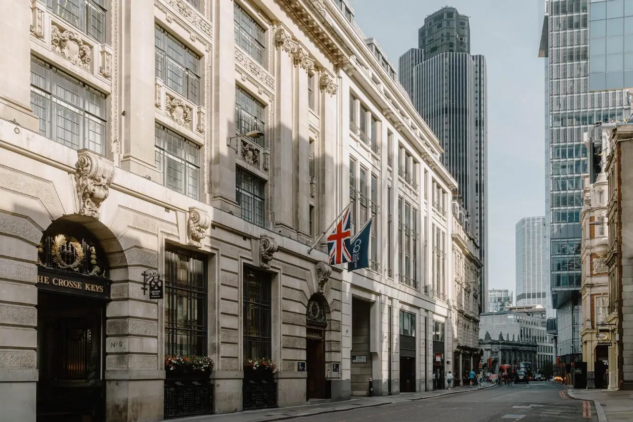 Street-level view of the stone facade of The Crosse Keys hotel, with a Union Jack and a blue flag hanging from the building and tall glass towers rising behind it.