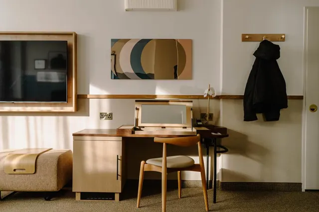 A hotel room with a wall-mounted television, a wooden desk with a mirror and lamp, a wooden chair, an upholstered bench, and a black jacket hanging on a wall hook.