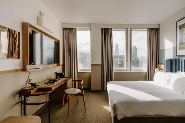 A bedroom with a made bed, a wall-mounted television, a desk and chair, and windows showing a city skyline with a tall glass tower.