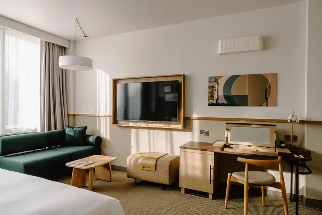 A hotel bedroom with a green sofa, a wall-mounted television, a desk with a mirror and a wooden chair, and a wall-mounted air conditioning unit.