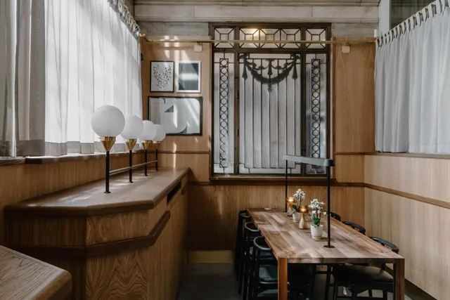A dining room with wood-panelled walls, a long wooden table with black chairs, a row of globe lamps on a counter, and an ornate window with white curtains.