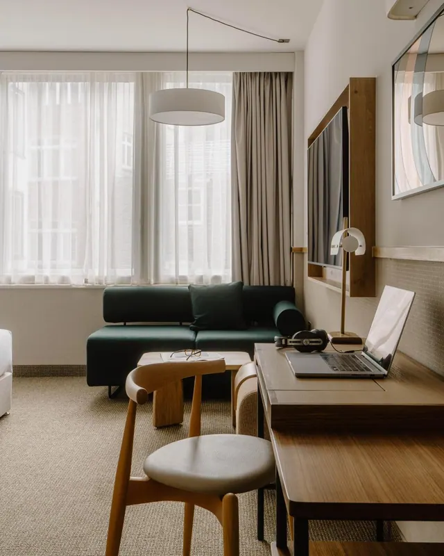 A hotel room living area with a green sofa, a wooden desk holding a laptop and headphones, a wall-mounted television, and a window with sheer curtains looking onto a brick building.