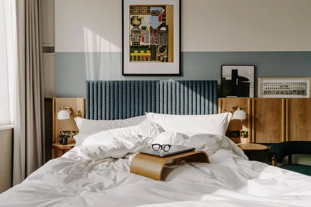 A made double bed with white linen, a blue tufted headboard, wooden bedside tables with wall lamps, and a laptop and glasses on a wooden bed tray.