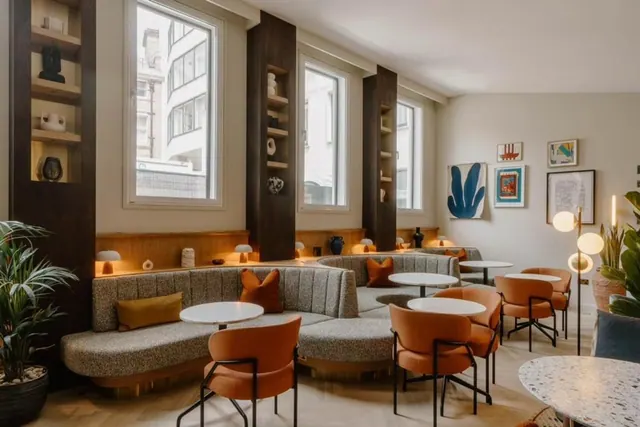 A lounge area with curved upholstered banquette seating, orange chairs and round tables beside tall windows and built-in shelving.