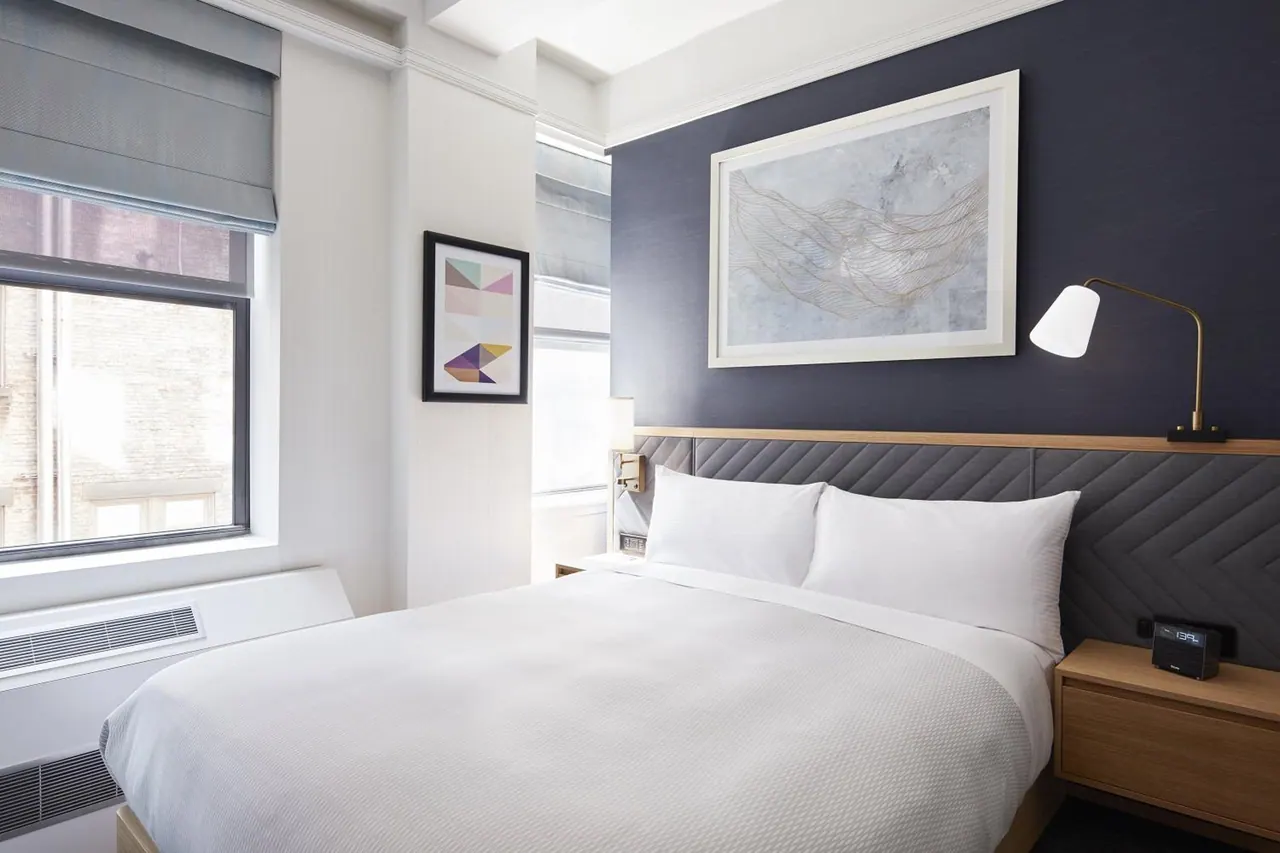 A made bed with white bedding sits beside a window, with a framed artwork above the headboard and a wall air conditioner unit below the window.