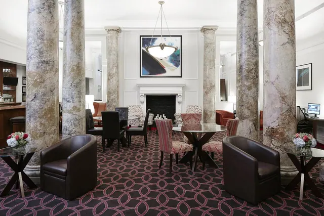 A lobby lounge seating area with marble columns, leather armchairs, upholstered chairs, round glass tables and a fireplace beneath a framed painting.