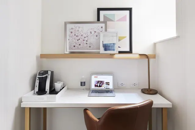 A white desk with a brown leather chair, a coffee machine on a tray, a laptop, a desk lamp and framed art on a wooden shelf above.