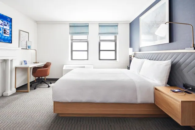 A bedroom with a king bed under white linens, a wooden nightstand, a work desk with a brown chair, and a wall-mounted television.