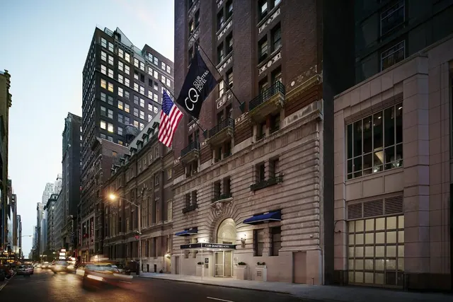 The exterior of the Club Quarters Hotel on a city street at dusk, with an American flag and a hotel flag hanging from the brick facade.