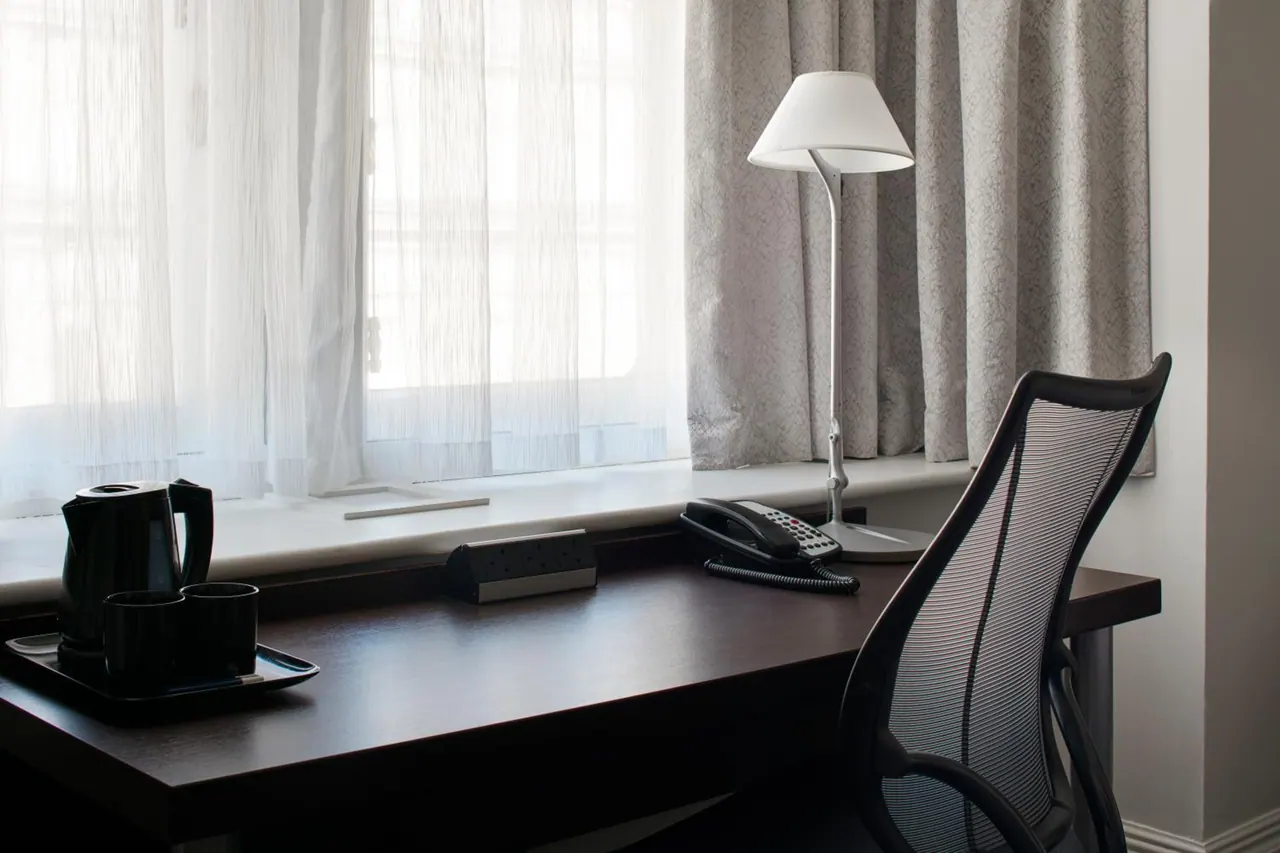 A dark wooden desk by a window with sheer and grey curtains holds a black kettle and cups on a tray, a corded telephone, a power socket strip, and a white desk lamp beside a mesh office chair.