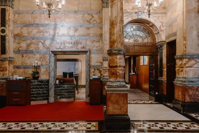 A marble-walled lobby with a red carpet, a reception desk with a monitor, chandeliers, and a Club Quarters sign above an arched doorway.
