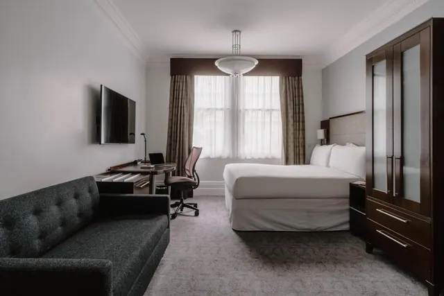 A hotel bedroom with a made double bed, a grey tufted sofa, a wall-mounted television, a desk with a chair, and a tall wooden wardrobe.