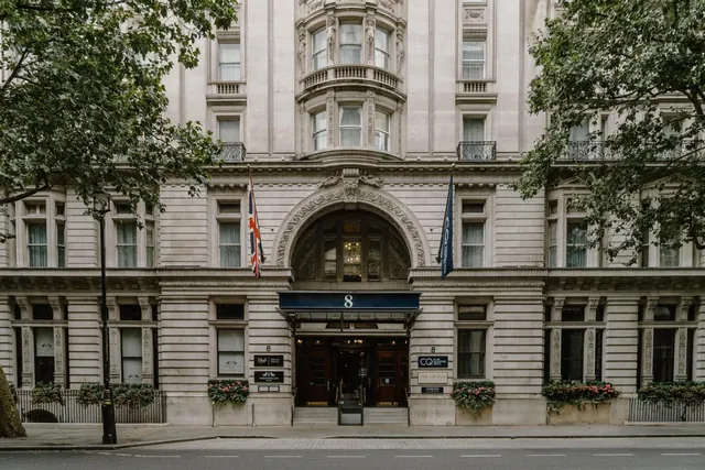 The stone front of a hotel building with an arched central entrance, a blue awning marked 8, flags, flower boxes and trees along the street.