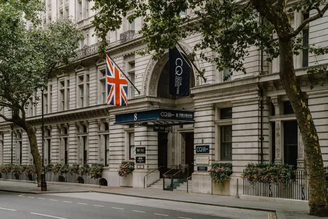 The stone front of a hotel building with a Union Jack flag, a navy canopy reading 8 The Grand, and a navy banner on the facade, seen from the street past tree branches.