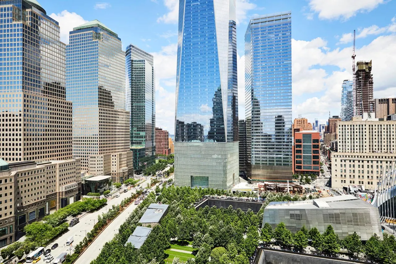 Elevated view of One World Trade Center and surrounding Lower Manhattan skyscrapers, with trees of the memorial plaza in the foreground.