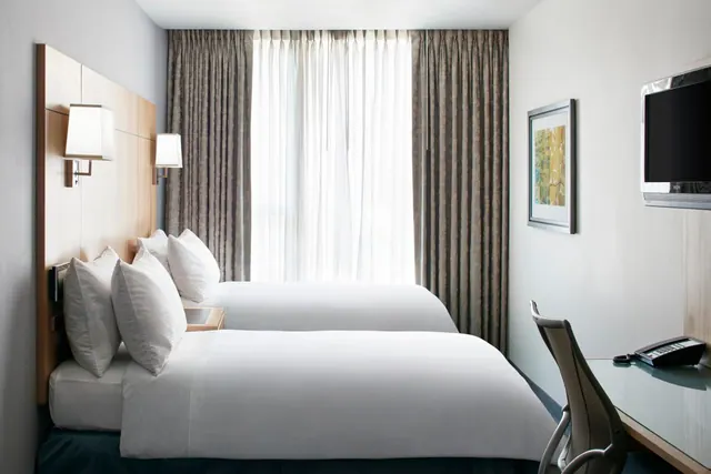 A hotel bedroom with two beds made up in white linen, sheer curtains at the window, a wall-mounted television and a desk with a chair on the right.