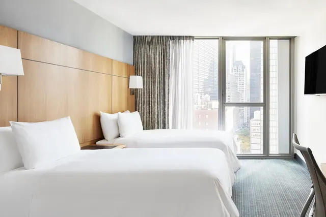 Two made-up beds face floor-to-ceiling windows with a view of city buildings, beside a wall-mounted television and a desk chair.