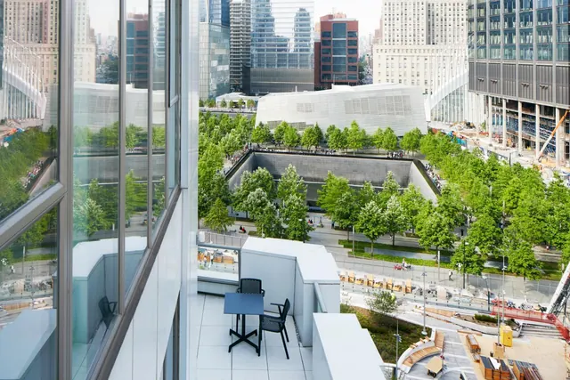 A balcony with a small table and chairs overlooks the sunken National September 11 Memorial pools, tree-lined plaza, and surrounding skyscrapers.