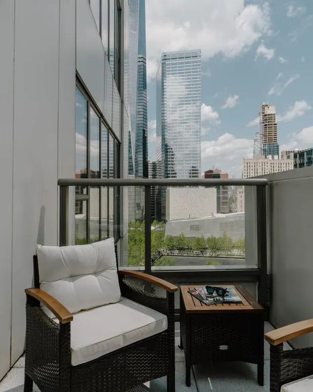 A private balcony with two wicker armchairs with white cushions and a small side table holding a magazine and sunglasses, looking out over high-rise towers and a low white building with trees below.