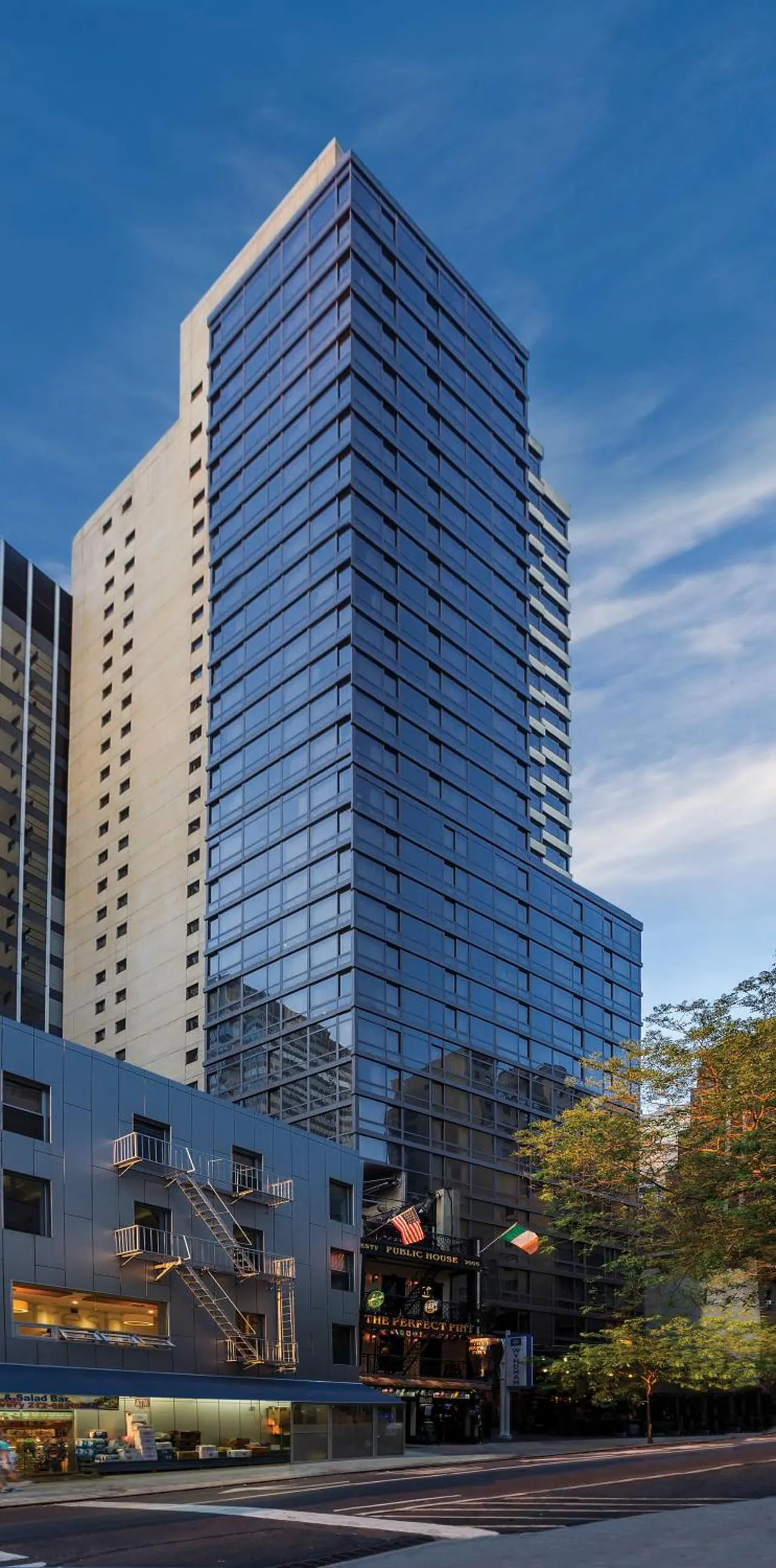 Exterior view of a tall glass and light-stone tower under a blue sky, with a street-level bar and a lower building with fire escapes in front.