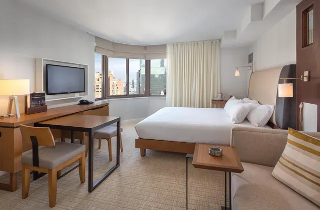 A hotel bedroom with a king bed, a work desk, a wall-mounted television and a sofa, with a bay window looking out over city buildings.