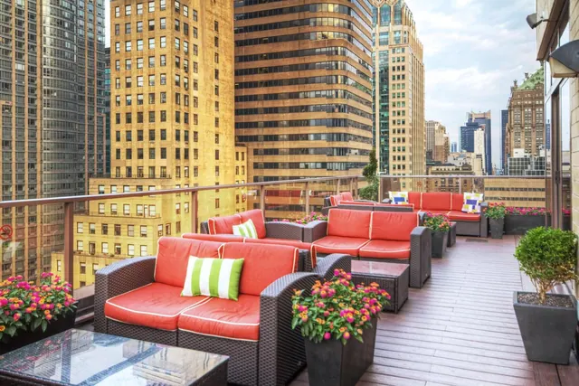 A rooftop terrace with red wicker sofas, striped cushions and potted flowers overlooks tall city buildings.