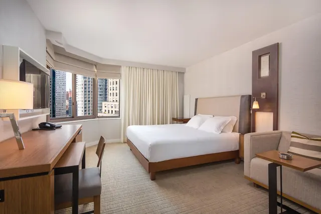 A hotel bedroom with a made double bed, a work desk, an upholstered armchair, and a window looking out onto city buildings.