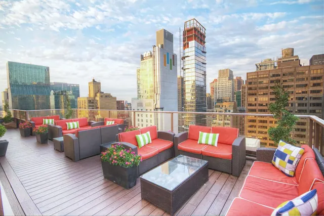 A rooftop terrace with red-cushioned wicker sofas, a glass coffee table and potted plants, with city skyscrapers visible beyond the railing.